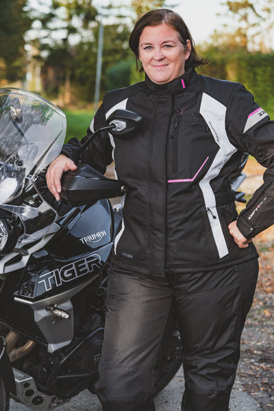 A smiling woman standing by her Triumph Tiger motorcycle wearing Difi textile women's motorcycle Nexia jacket and Cyclone pants