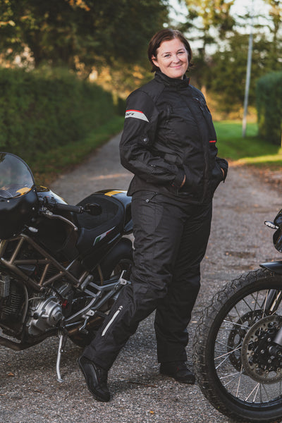 A smiling woman standing by motorcycles wearing DIFI motorcycle black Cyclone pants and Nexia jacket